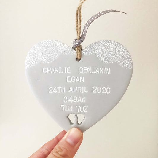 Personalised baby keepsake gift, grey clay hanging heart with baby feet cut out at the bottom of the heart and white lace edge to the top, the heart is personalised with the baby’s name, date of birth, weight and time