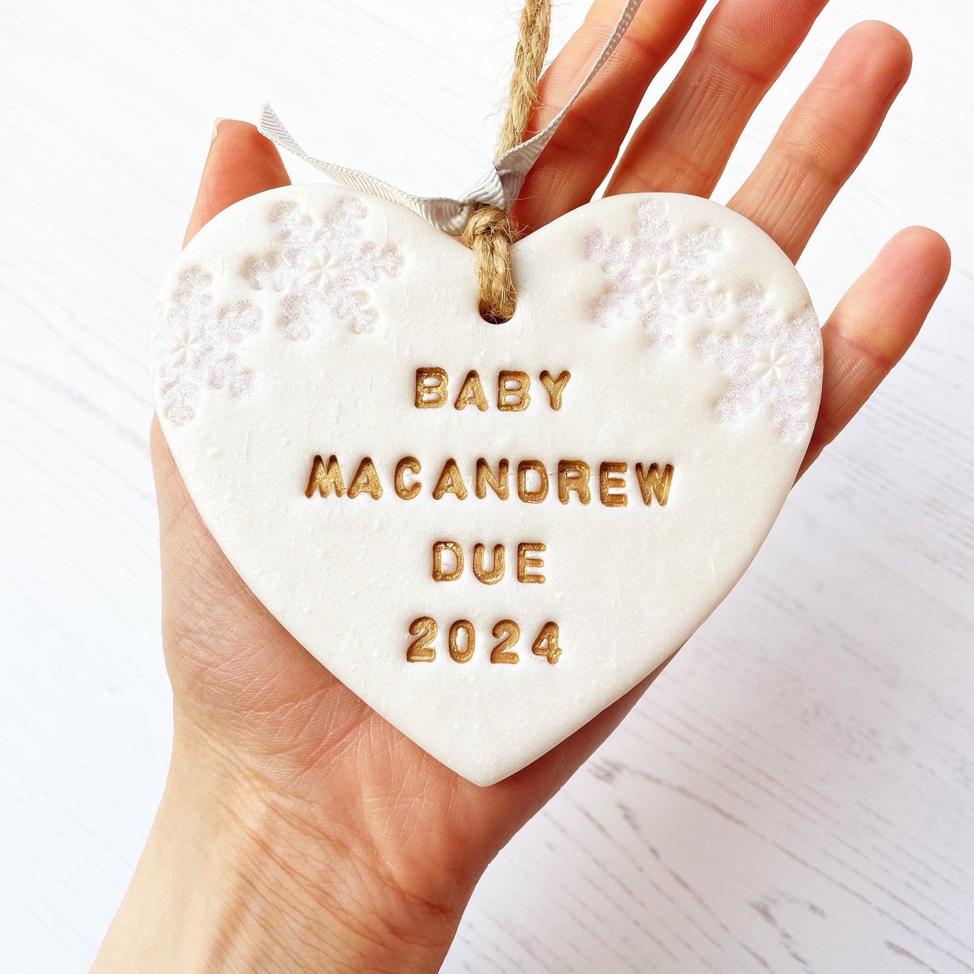 Personalised baby's first Christmas heart ornament, pearlised white clay with BABY MACANDREW DUE 2024 painted gold, decorated with 2 iridescent glitter snowflakes on either side of the top of the heart