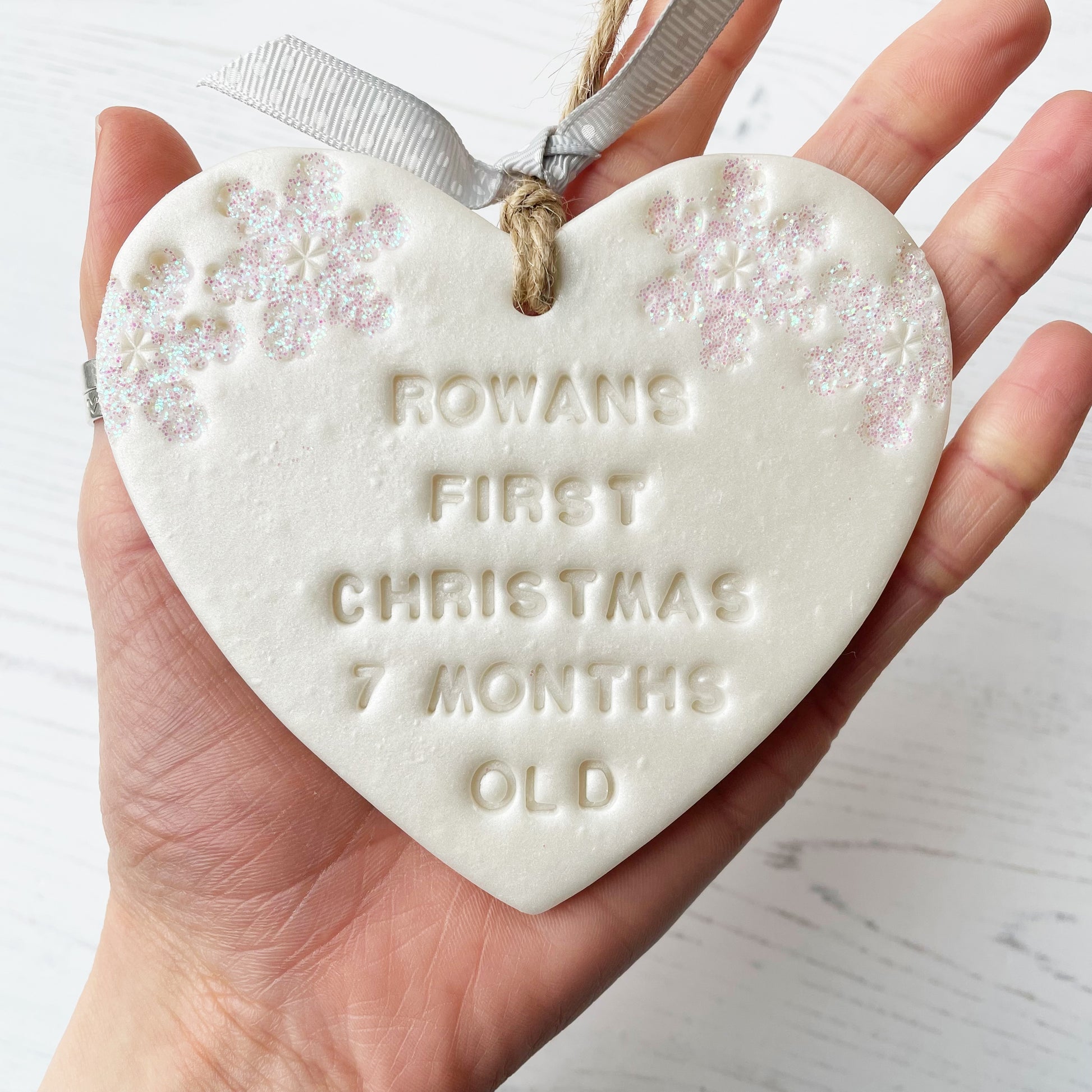 Personalised baby’s first Christmas heart ornament, pearlised white clay with ROWANS FIRST CHRISTMAS 7 MONTHS OLD (text not painted), decorated with 2 iridescent glitter snowflakes on either side of the top of the heart