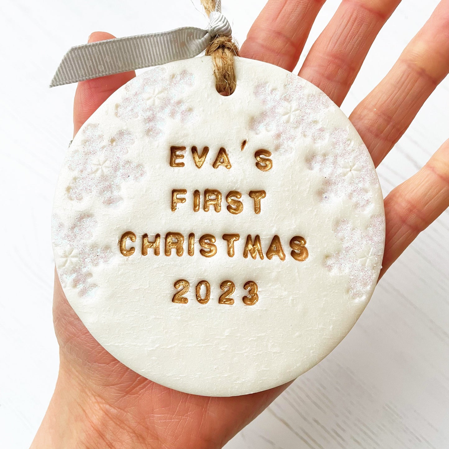 Personalised baby’s first Christmas bauble ornament, pearlised white clay round personalised with Eva’s first Christmas 2023 in gold paint, decorated with 3 iridescent glitter snowflakes on either side of the bauble