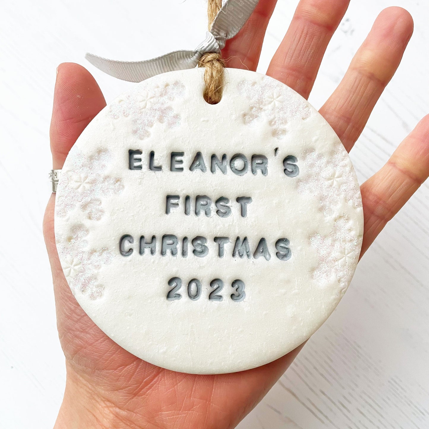 Personalised baby’s first Christmas bauble ornament, pearlised white clay round personalised with Eleanor’s first Christmas 2023 in grey paint, decorated with 3 iridescent glitter snowflakes on either side of the bauble