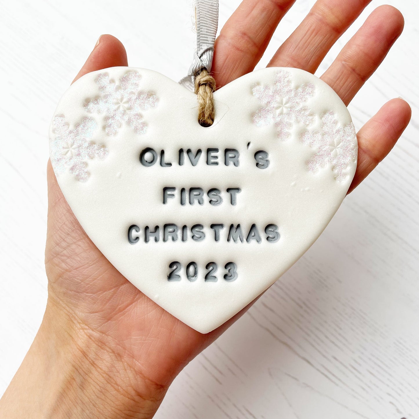 Personalised baby's first Christmas heart ornament, pearlised white clay with OLIVER'S FIRST CHRISTMAS 2023 painted grey, decorated with 2 iridescent glitter snowflakes on either side of the top of the heart