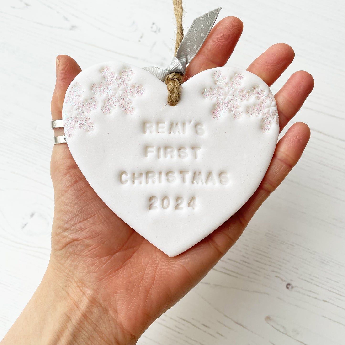 Personalised baby’s first Christmas heart ornament, glitter white clay with REMI’S FIRST CHRISTMAS 2024 (text not painted), decorated with 2 iridescent glitter snowflakes on either side of the top of the heart