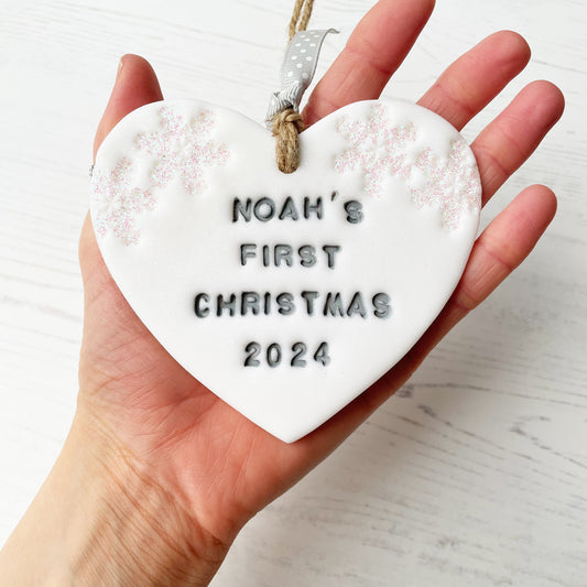 Personalised baby’s first Christmas heart ornament, glitter white clay with NOAH’S FIRST CHRISTMAS 2024 painted grey, decorated with 2 iridescent glitter snowflakes on either side of the top of the heart