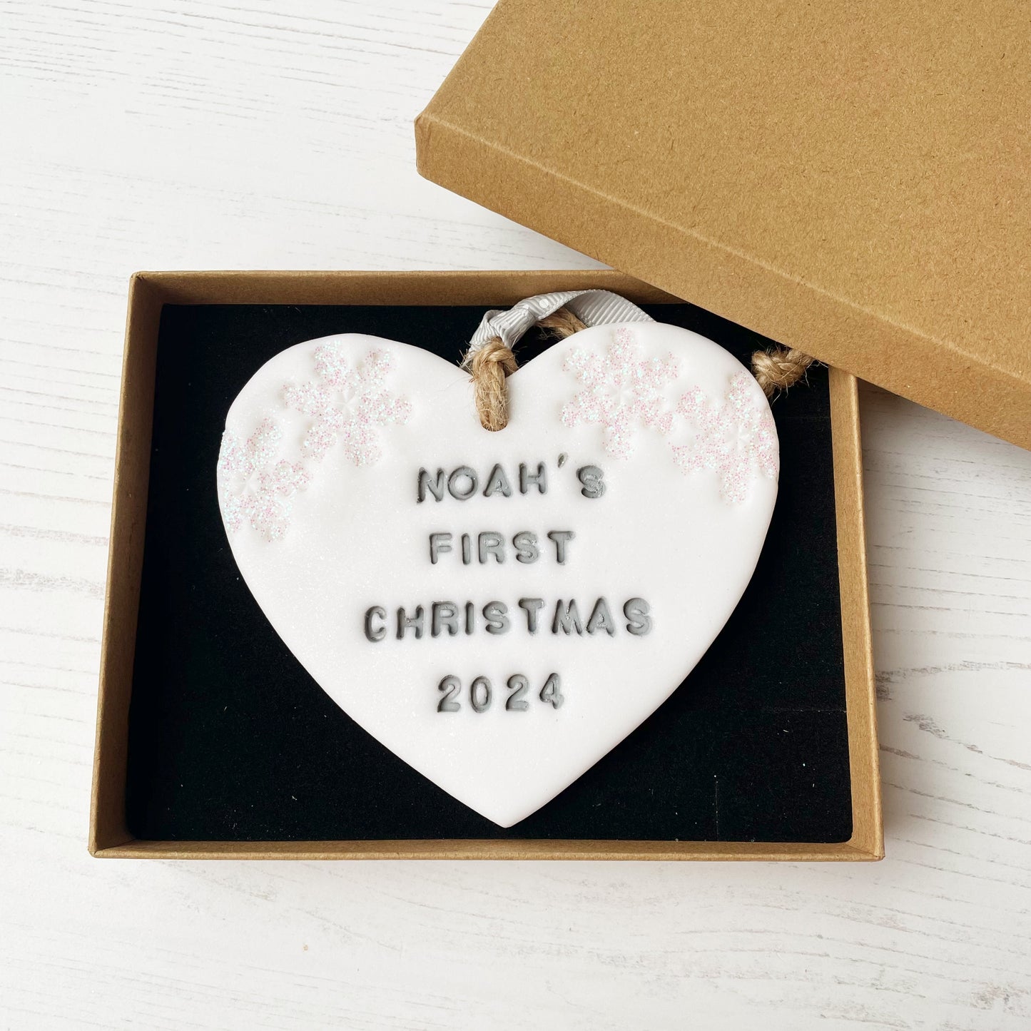 Personalised baby’s first Christmas heart ornament, glitter white clay with NOAH’S FIRST CHRISTMAS 2024 painted grey, decorated with 2 iridescent glitter snowflakes on either side of the top of the heart