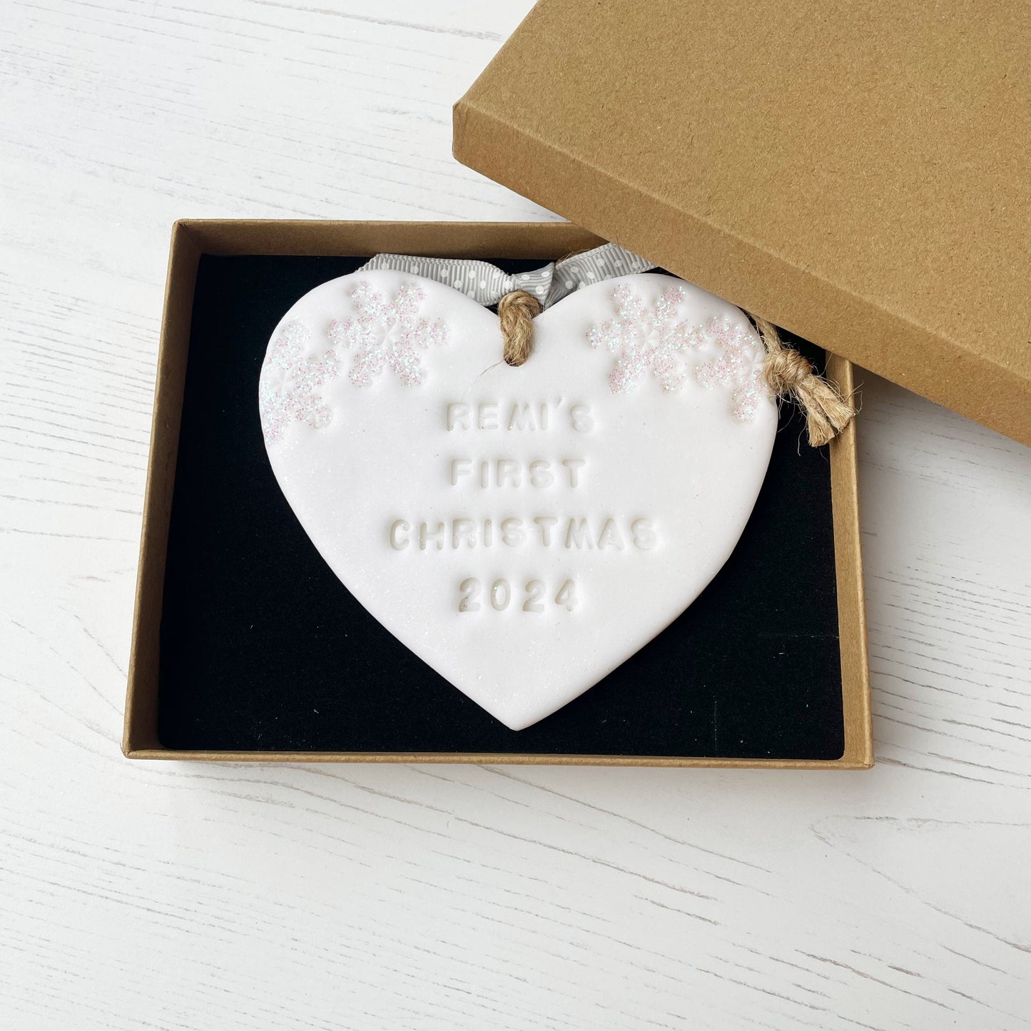 Personalised baby’s first Christmas heart ornament, glitter white clay with REMI’S FIRST CHRISTMAS 2024 (text not painted), decorated with 2 iridescent glitter snowflakes on either side of the top of the heart