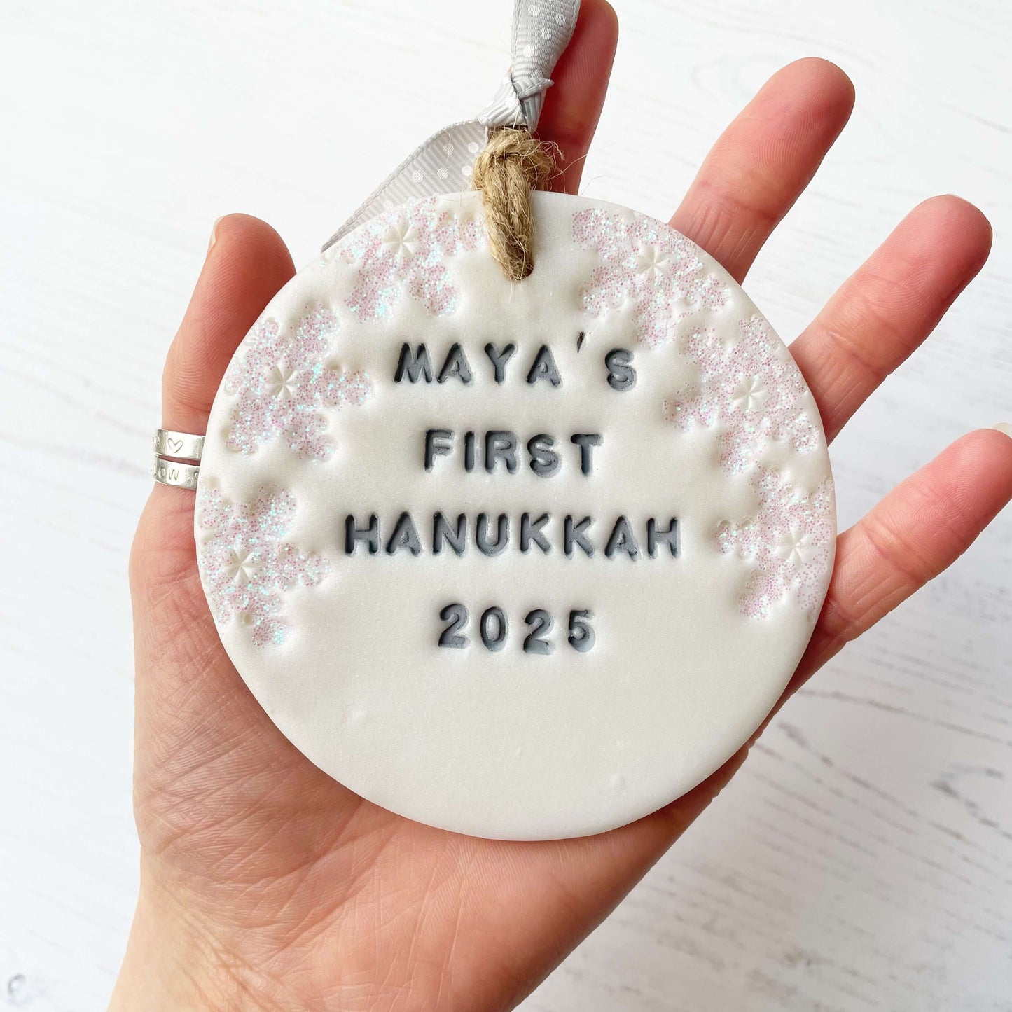 Personalised baby's first Hanukkah bauble ornament, pearlised white round clay with MAYA’S FIRST HANUKKAH 2025 in grey paint, decorated with 3 iridescent glitter snowflakes on either side of the bauble