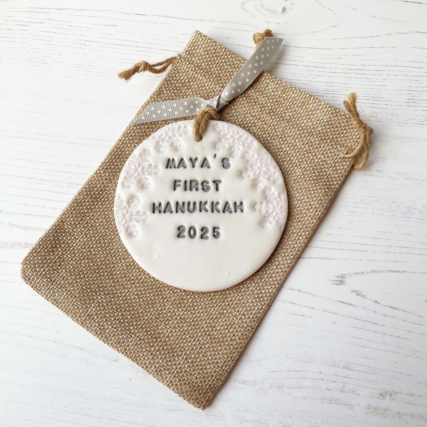 Personalised baby's first Hanukkah bauble ornament, pearlised white round clay with MAYA’S FIRST HANUKKAH 2025 in grey paint, decorated with 3 iridescent glitter snowflakes on either side of the bauble
