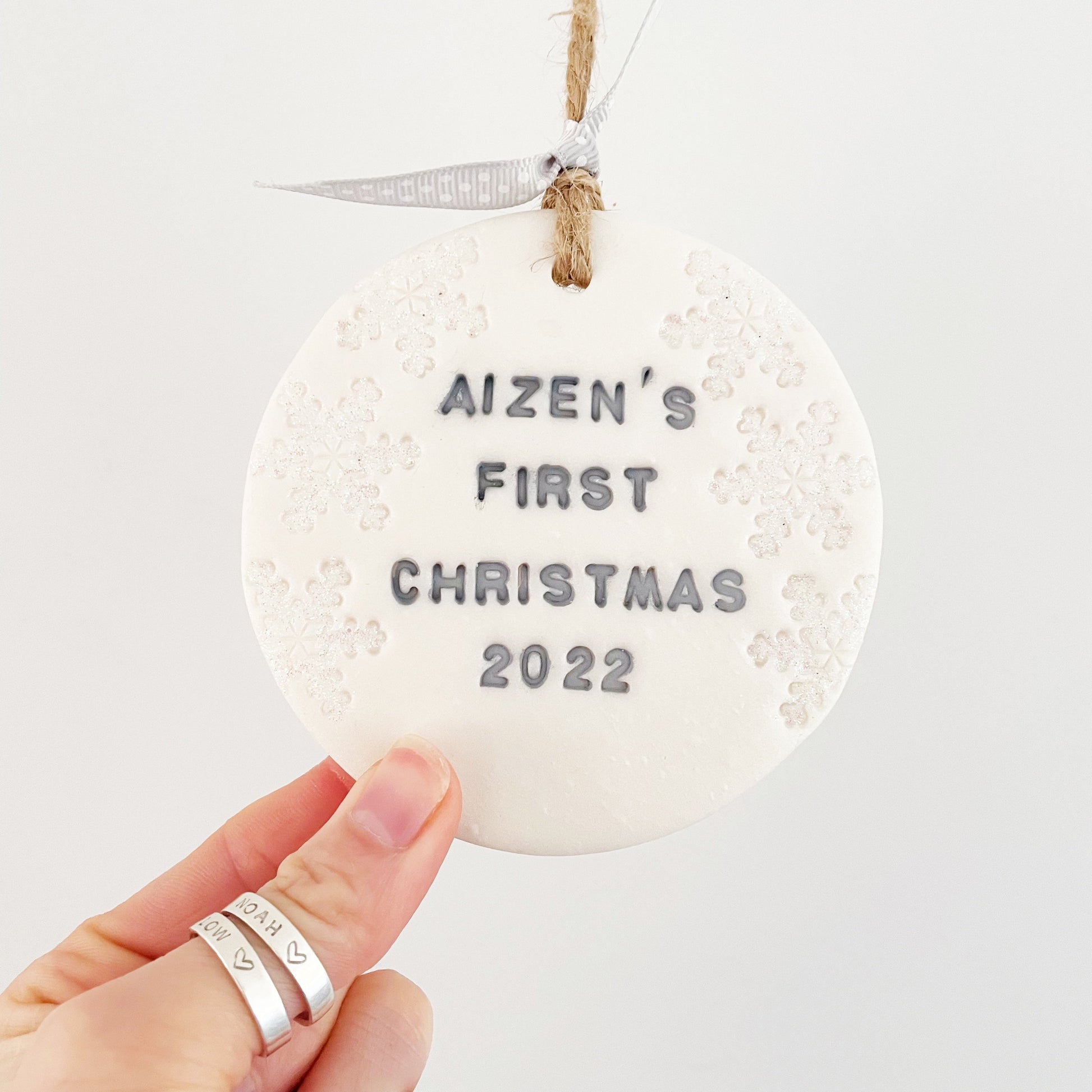 Personalised baby's first Christmas bauble ornament, pearlised white round clay with AIZEN’S FIRST CHRISTMAS 2022 in grey paint, decorated with 3 iridescent glitter snowflakes on either side of the bauble