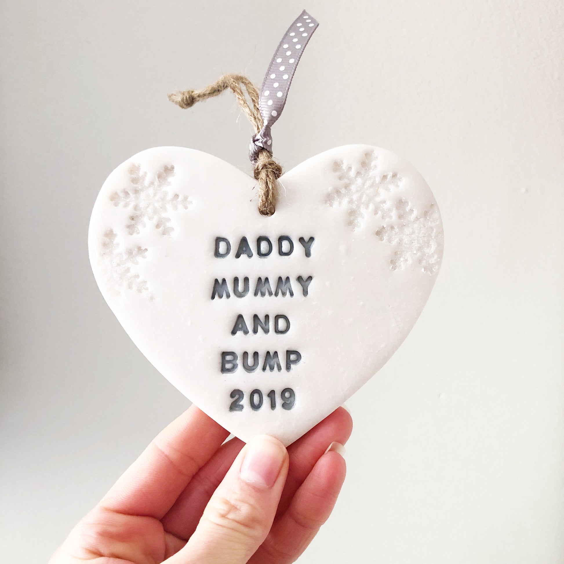 Personalised bump's first Christmas heart ornament, pearlised white clay with DADDY MUMMY AND BUMP 2019 painted grey, decorated with 2 iridescent glitter snowflakes on either side of the top of the heart