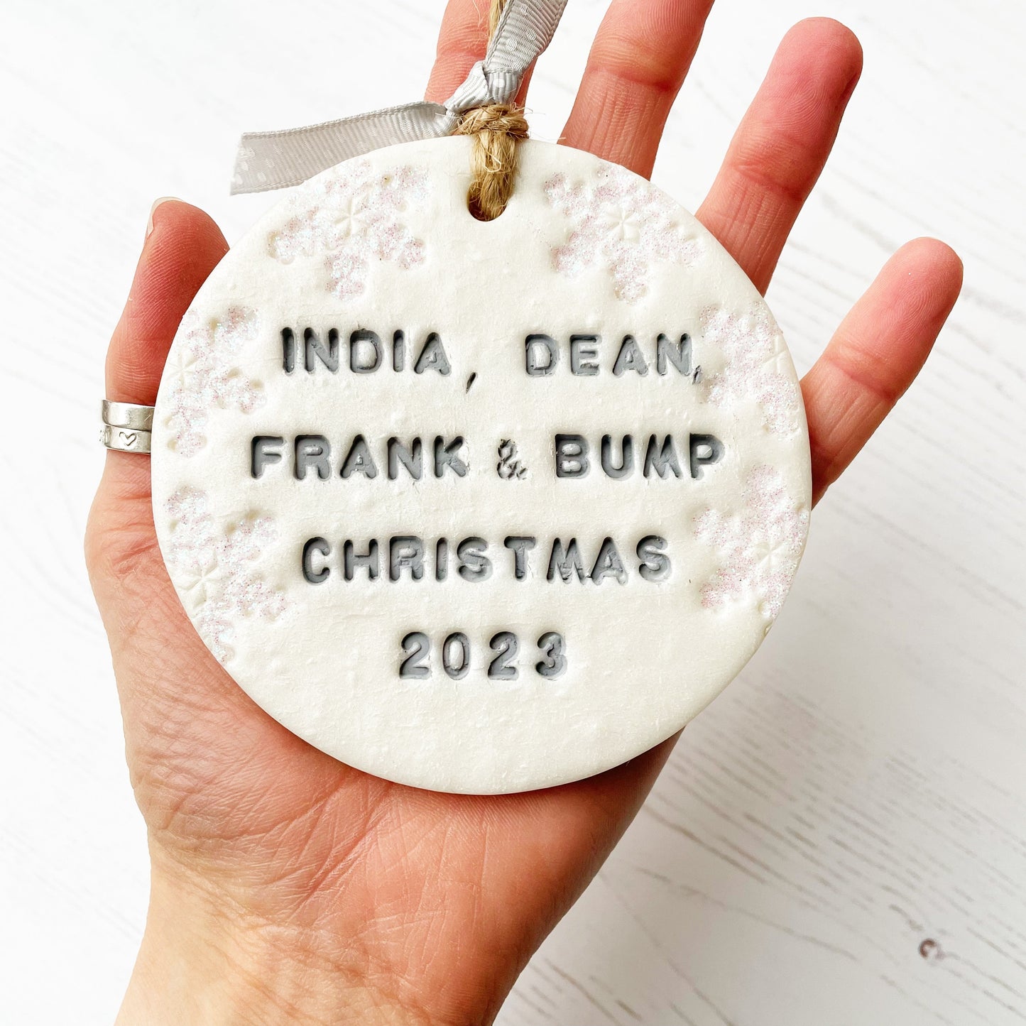 Personalised bump’s first Christmas bauble ornament, pearlised white round clay with INDIA, DEAN, FRANK & BUMP CHRISTMAS 2023 in grey paint, decorated with 3 iridescent glitter snowflakes on either side of the bauble