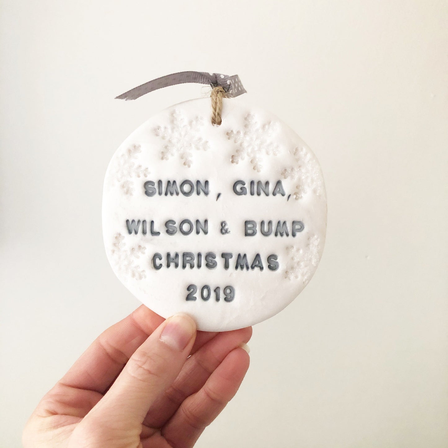 Personalised baby's first Christmas bauble ornament, pearlised white round clay with SIMON, GINA, WILSON & BUMP CHRISTMAS 2019 in grey paint, decorated with 3 iridescent glitter snowflakes on either side of the bauble