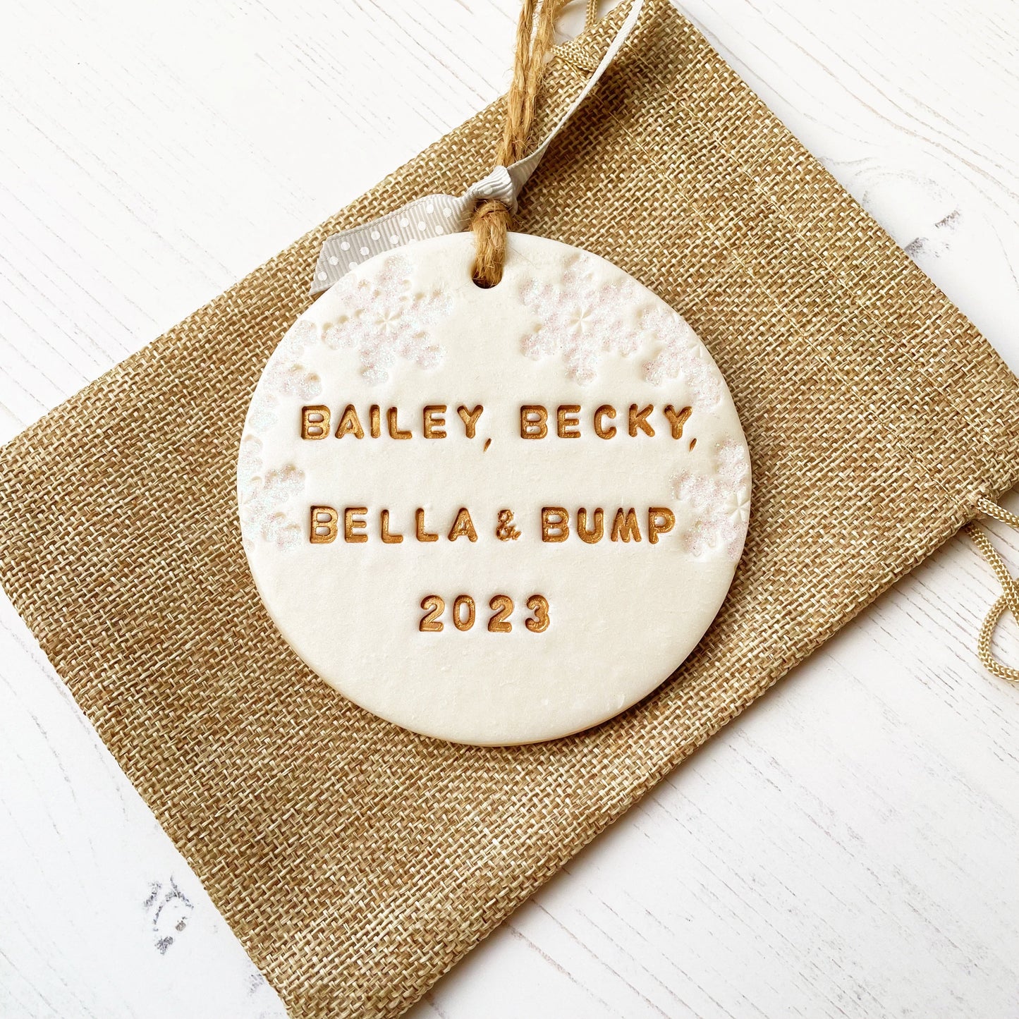 Personalised bump’s first Christmas bauble ornament, pearlised white round clay with BAILEY, BECKY, BELLA & BUMP 2023 in gold paint, decorated with 3 iridescent glitter snowflakes on either side of the bauble