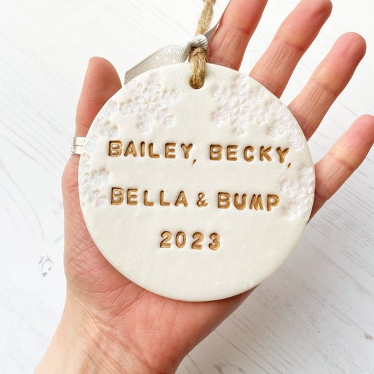 Personalised bump’s first Christmas bauble ornament, pearlised white round clay with BAILEY, BECKY, BELLA & BUMP 2023 in gold paint, decorated with 3 iridescent glitter snowflakes on either side of the bauble