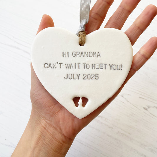 Pearlised white clay hanging heart with baby feet cut out of the bottom and grey personalisation, the heart is personalised with HI GRANDMA CAN’T WAIT TO MEET YOU! JULY 2025