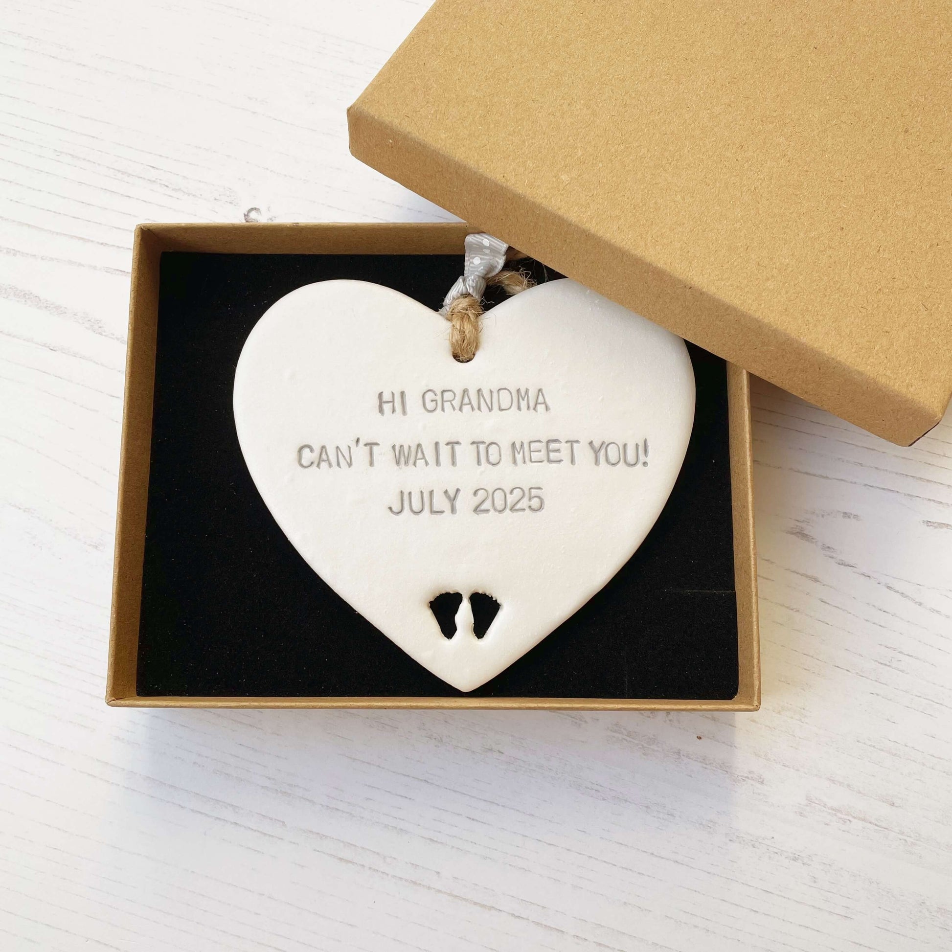 Pearlised white clay hanging heart with baby feet cut out of the bottom and grey personalisation, the heart is personalised with HI GRANDMA CAN’T WAIT TO MEET YOU! JULY 2025