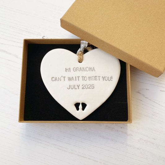 Pearlised white clay hanging heart with baby feet cut out of the bottom and grey personalisation, the heart is personalised with HI GRANDMA CAN’T WAIT TO MEET YOU! JULY 2025