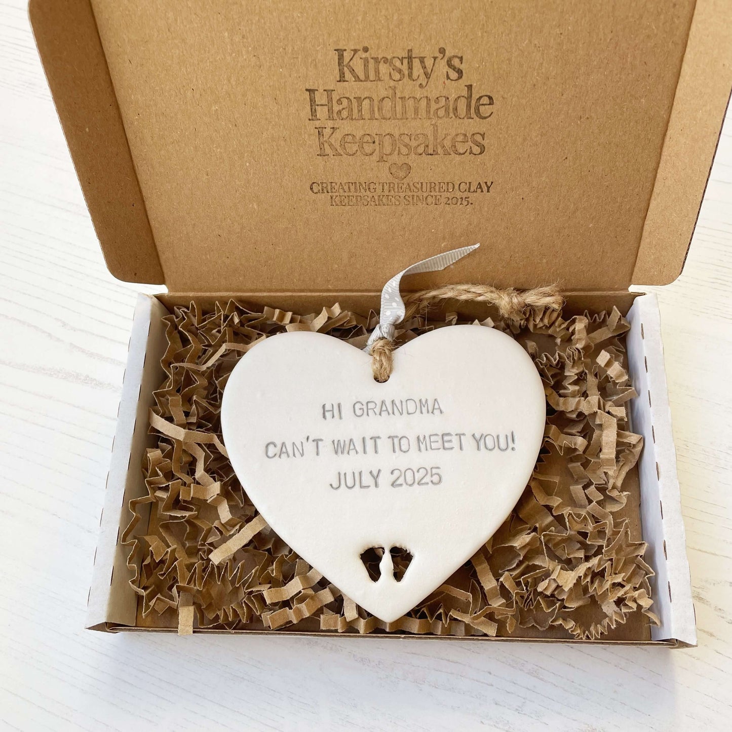 Pearlised white clay hanging heart with baby feet cut out of the bottom and grey personalisation, the heart is personalised with HI GRANDMA CAN’T WAIT TO MEET YOU! JULY 2025