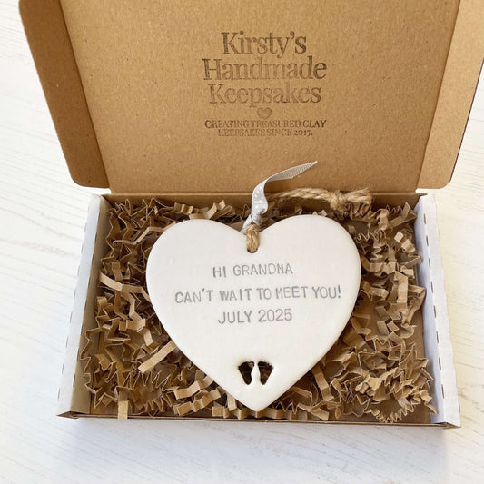 Pearlised white clay hanging heart with baby feet cut out of the bottom and grey personalisation, the heart is personalised with HI GRANDMA CAN’T WAIT TO MEET YOU! JULY 2025
