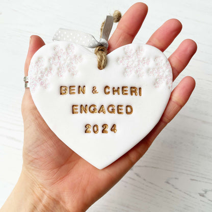 Personalised first Christmas engaged heart ornament, glitter white clay with BEN & CHERI ENGAGED 2024 painted gold, decorated with 2 iridescent glitter snowflakes on either side of the top of the heart