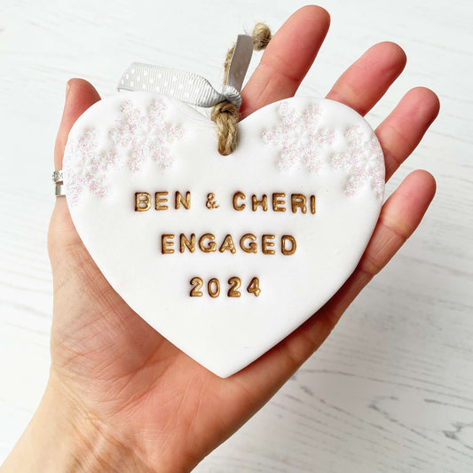 Personalised first Christmas engaged heart ornament, glitter white clay with BEN & CHERI ENGAGED 2024 painted gold, decorated with 2 iridescent glitter snowflakes on either side of the top of the heart