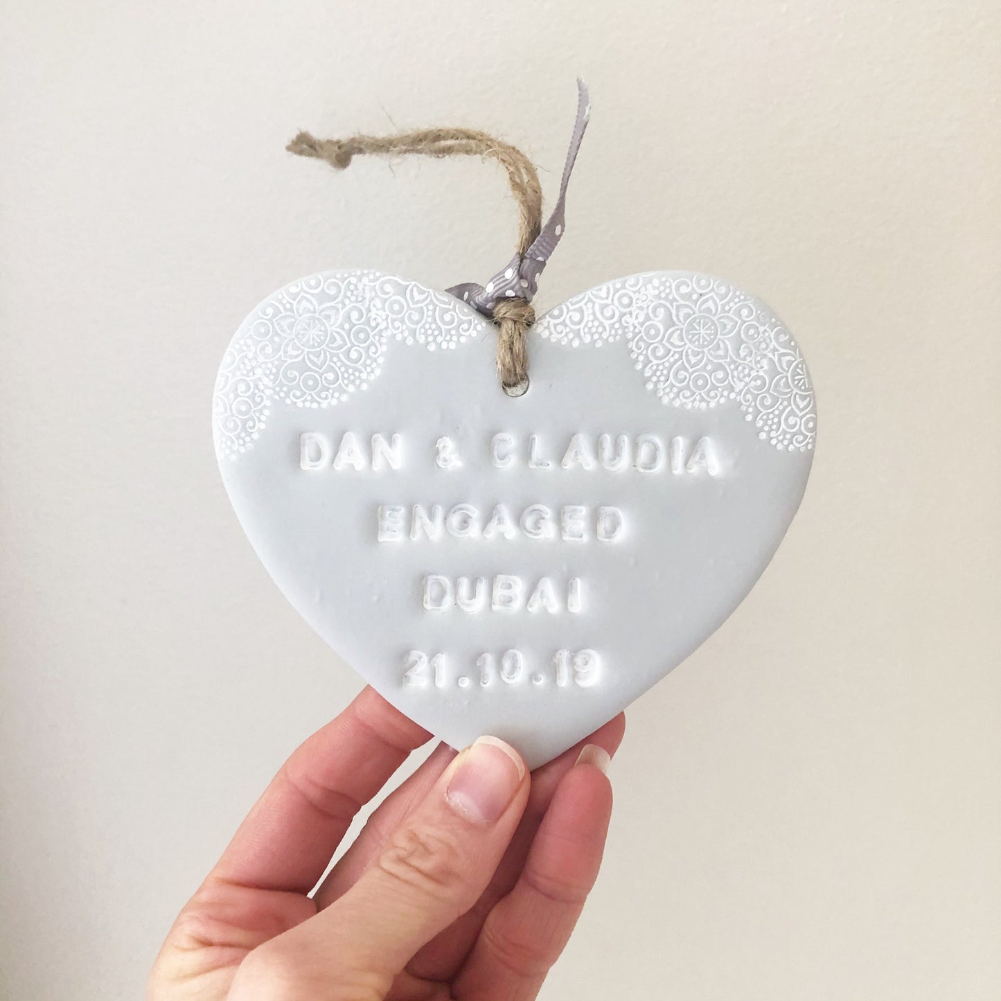 Personalised engaged abroad gift, grey clay heart with a white lace edge at the top of the heart with twine to hang, the heart is personalised with DAN & CLAUDIA ENGAGED DUBAI 21.10.19