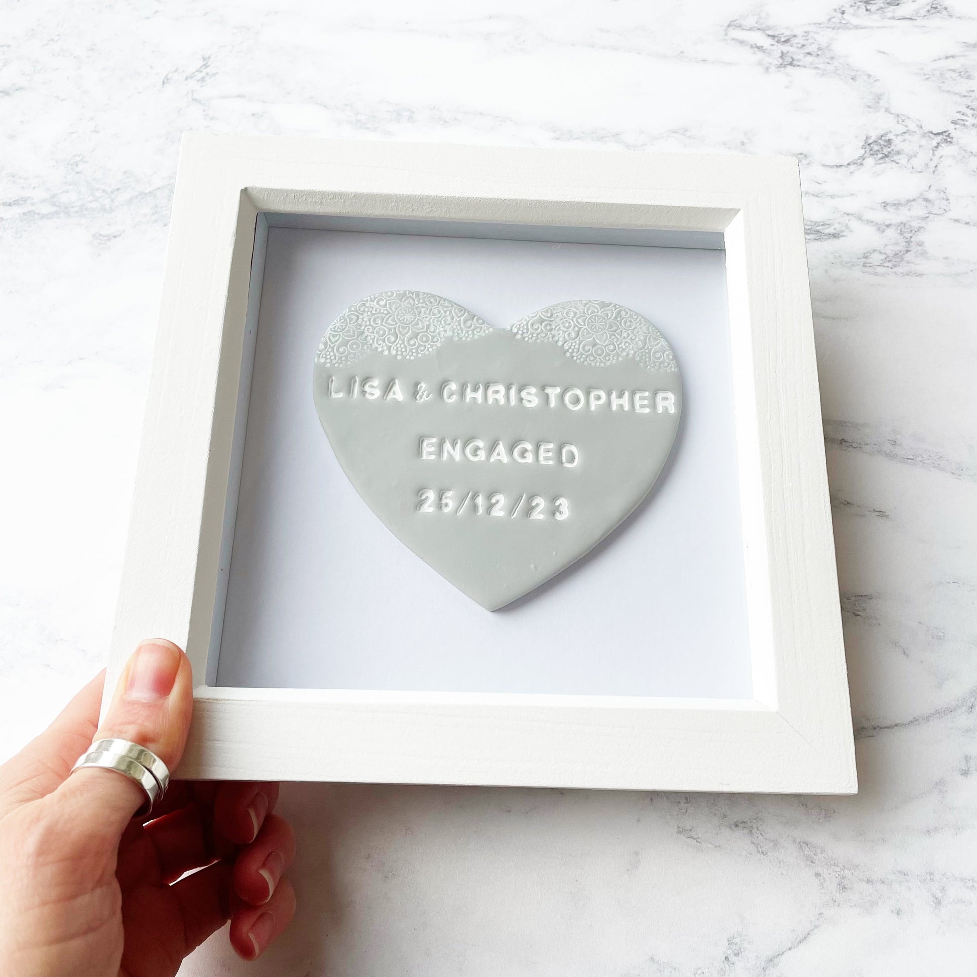 Personalised framed engagement gift, grey clay heart with a white lace edge at the top of the heart in a white box frame, the heart is personalised with LISA & CHRISTOPHER D 25/12/23