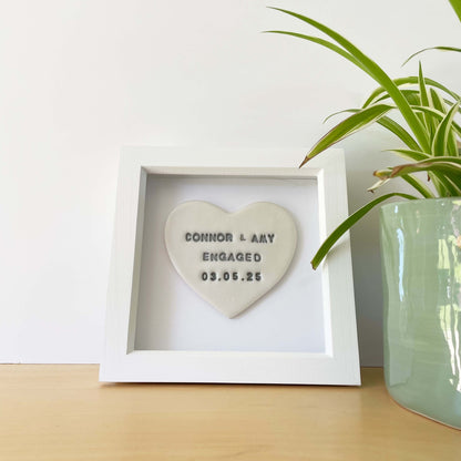 Personalised framed engagement gift, pearlised white clay heart in a white box frame, the heart is personalised in grey with CONNOR & AMY ENGAGED 03.05.25