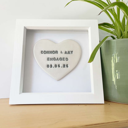 Personalised framed engagement gift, pearlised white clay heart in a white box frame, the heart is personalised in grey with CONNOR & AMY ENGAGED 03.05.25