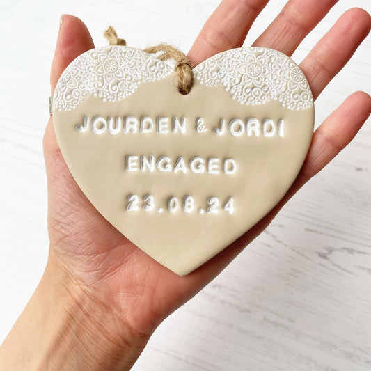 Personalised engagement gift, beige clay heart with a white lace edge at the top of the heart with jute twine for hanging, the heart is personalised with JOURDEN & JORDI ENGAGED 23.08.24