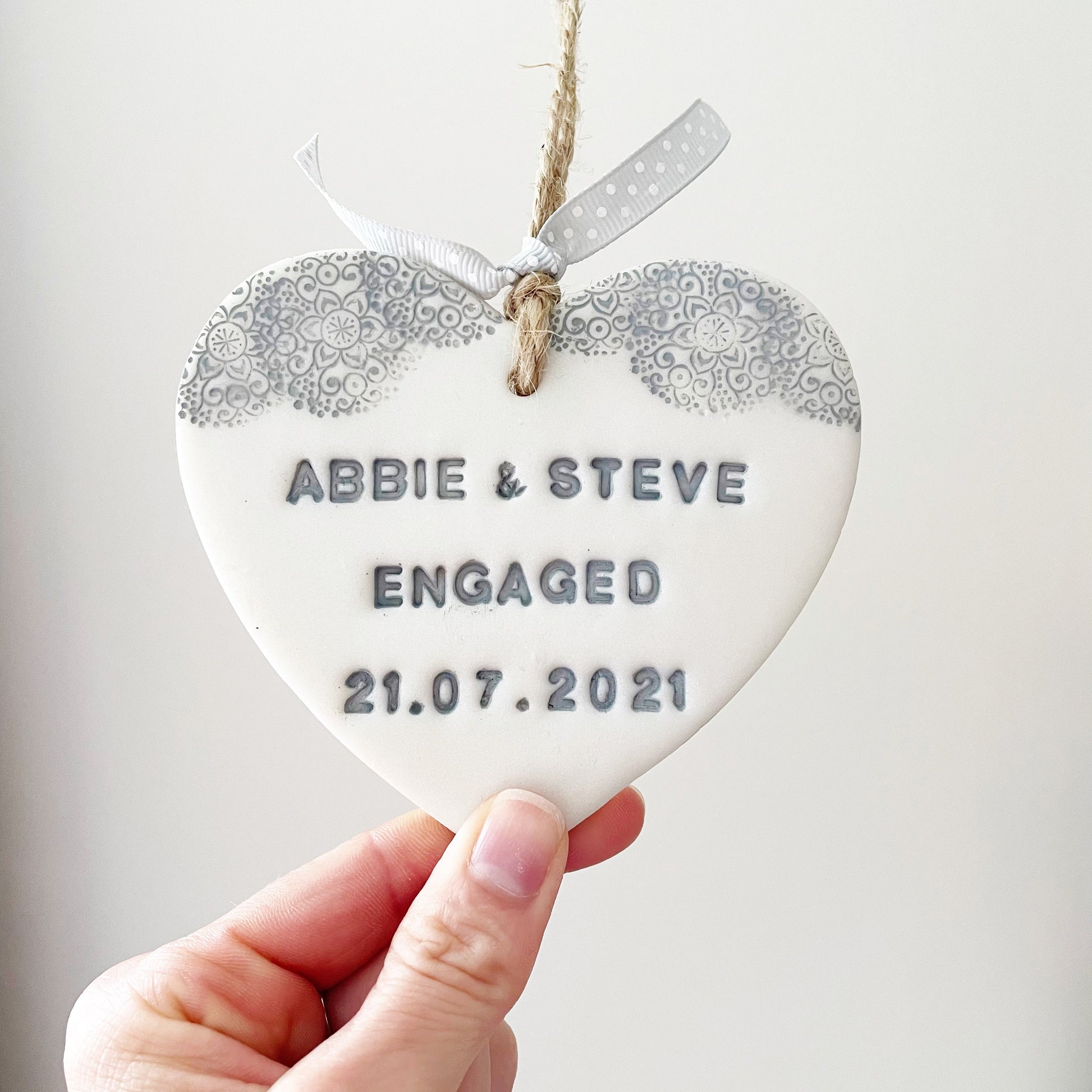 Personalised engagement gift, pearlised white clay heart with a grey lace edge at the top of the heart with jute twine for hanging, the heart is personalised with ABBIE & STEVE ENGAGED 21.07.2021