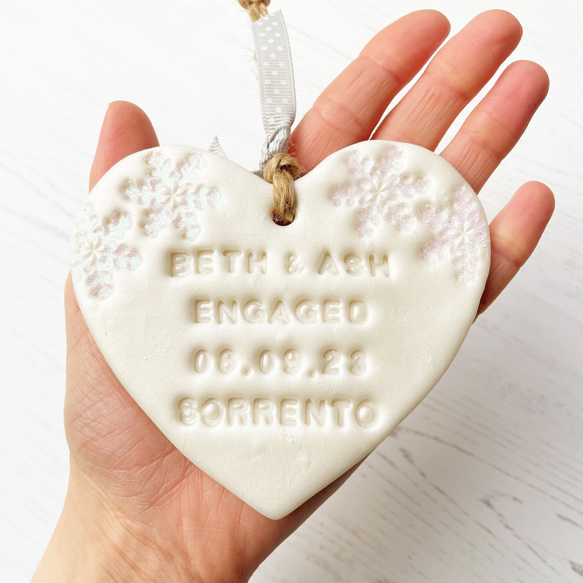 Personalised first Christmas engaged heart ornament, pearlised white clay with BETH & ASH ENGAGED 08.09.23 SORRENTO (text not painted), decorated with 2 iridescent glitter snowflakes on either side of the top of the heart