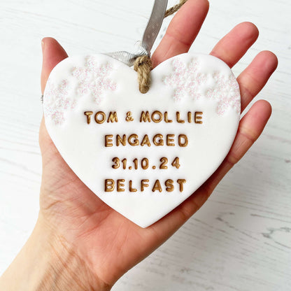 Personalised first Christmas engaged heart ornament, glitter white clay with TOM & MOLLIE ENGAGED 31.10.24 BELFAST painted gold, decorated with 2 iridescent glitter snowflakes on either side of the top of the heart