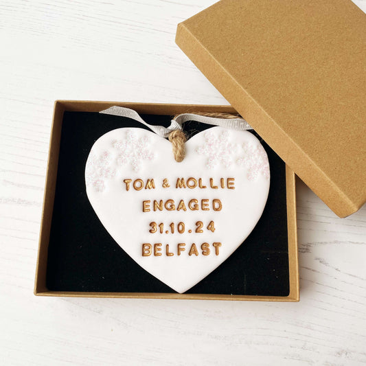 Personalised first Christmas engaged heart ornament, glitter white clay with TOM & MOLLIE ENGAGED 31.10.24 BELFAST painted gold, decorated with 2 iridescent glitter snowflakes on either side of the top of the heart
