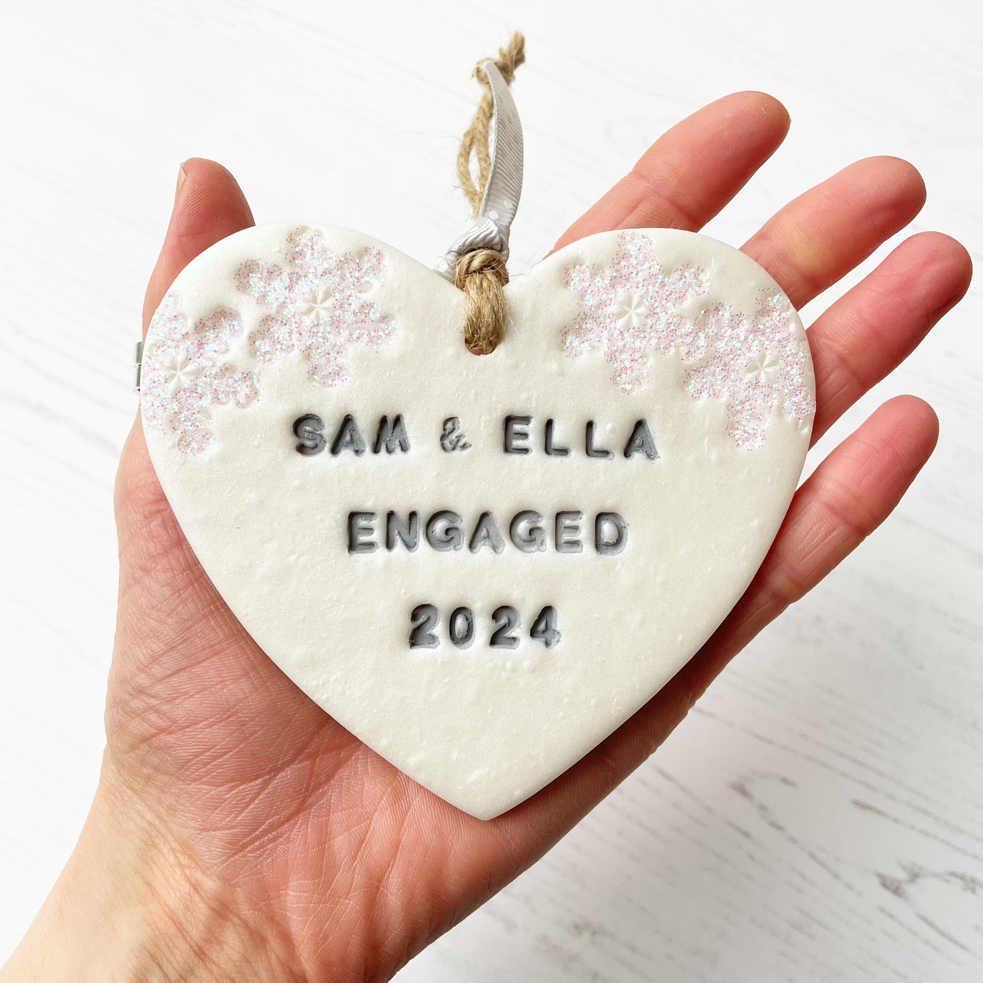 Personalised first Christmas engaged heart ornament, pearlised white clay with SAM & ELLA ENGAGED 2024 painted grey, decorated with 2 iridescent glitter snowflakes on either side of the top of the heart