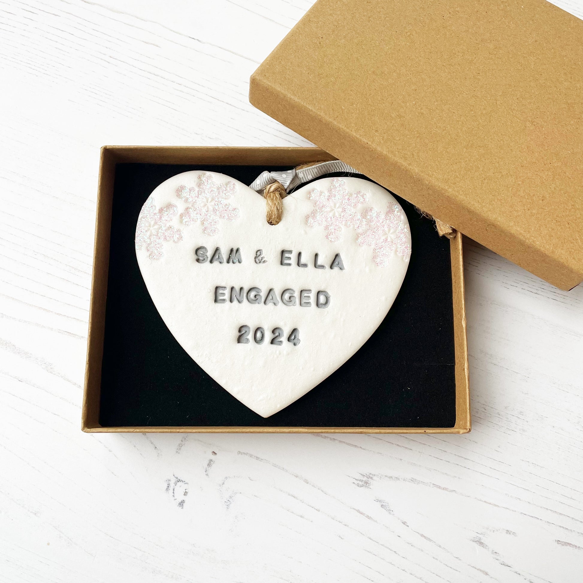 Personalised first Christmas engaged heart ornament, pearlised white clay with SAM & ELLA ENGAGED 2024 painted grey, decorated with 2 iridescent glitter snowflakes on either side of the top of the heart