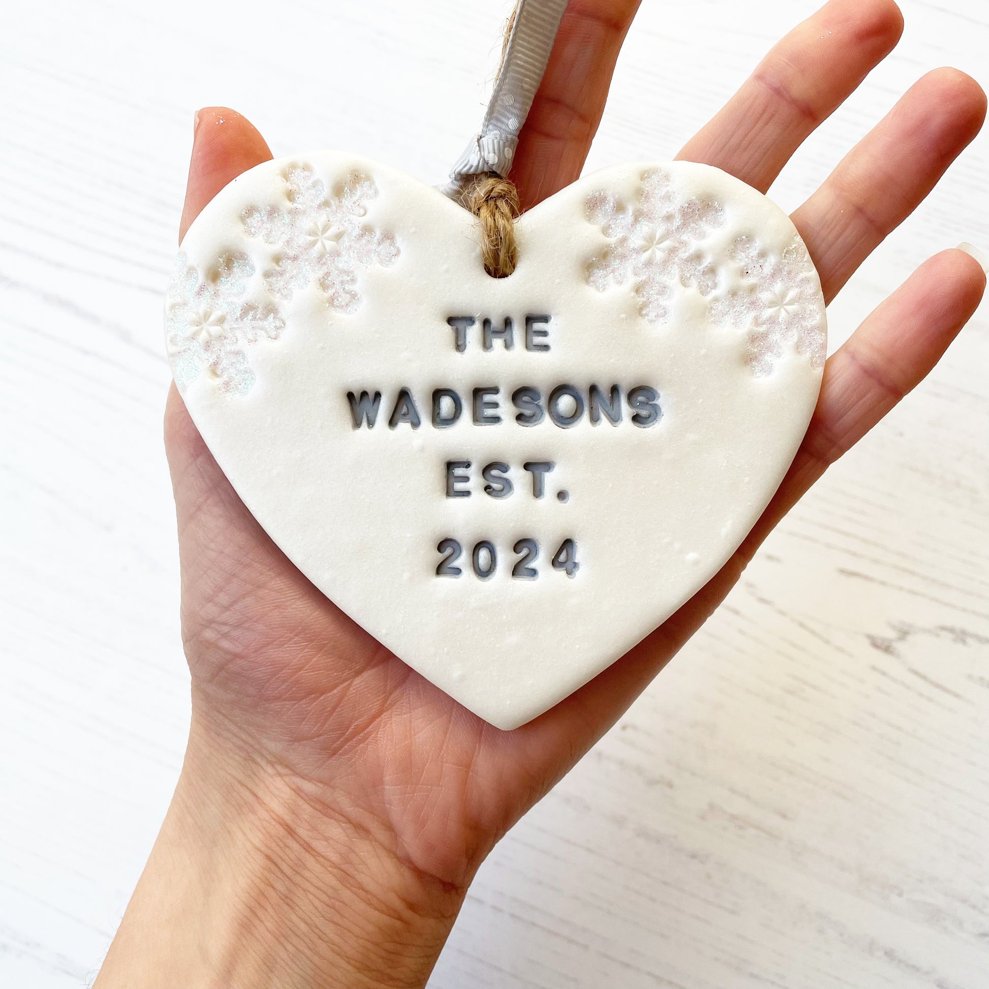 Personalised first Christmas married heart ornament, pearlised white clay personalised in grey with THE WADESONS EST. 2024, decorated with 2 iridescent glitter snowflakes on either side of the top of the heart