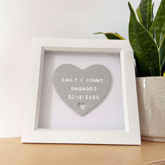 Personalised framed engagement gift, grey clay heart with a heart cut out of the bottom in a white box frame, the heart is personalised in white with EMILY & JONNY ENGAGED 20/12/2025