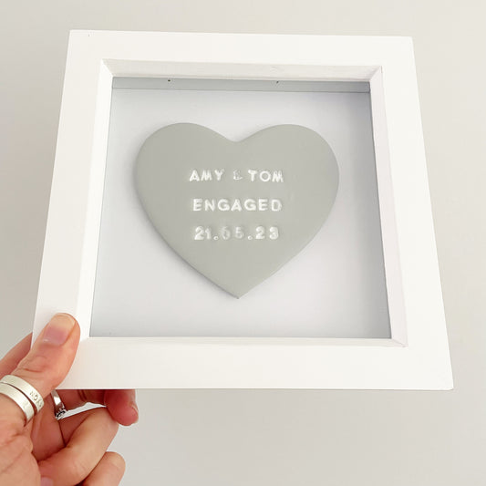 Personalised framed engagement gift, grey clay heart in a white box frame, the heart is personalised with AMY & TOM ENGAGED 21.05.23