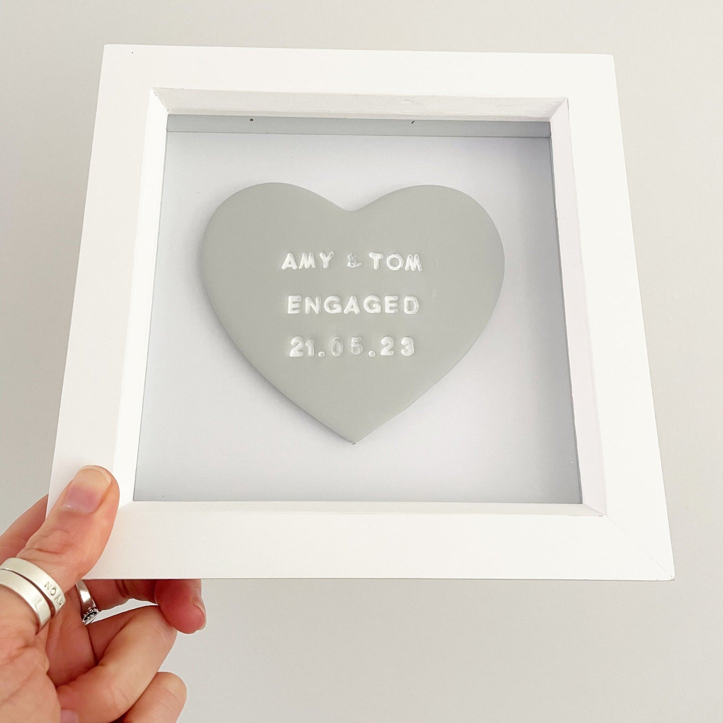 Personalised framed engagement gift, grey clay heart in a white box frame, the heart is personalised with AMY & TOM ENGAGED 21.05.23