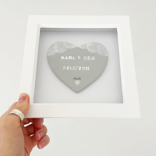 Personalised anniversary gift, grey clay heart with a white lace edge at the top of the heart and a heart cut out at the bottom in a white box frame, the heart is personalised with KARL & DEB 01/10/2011