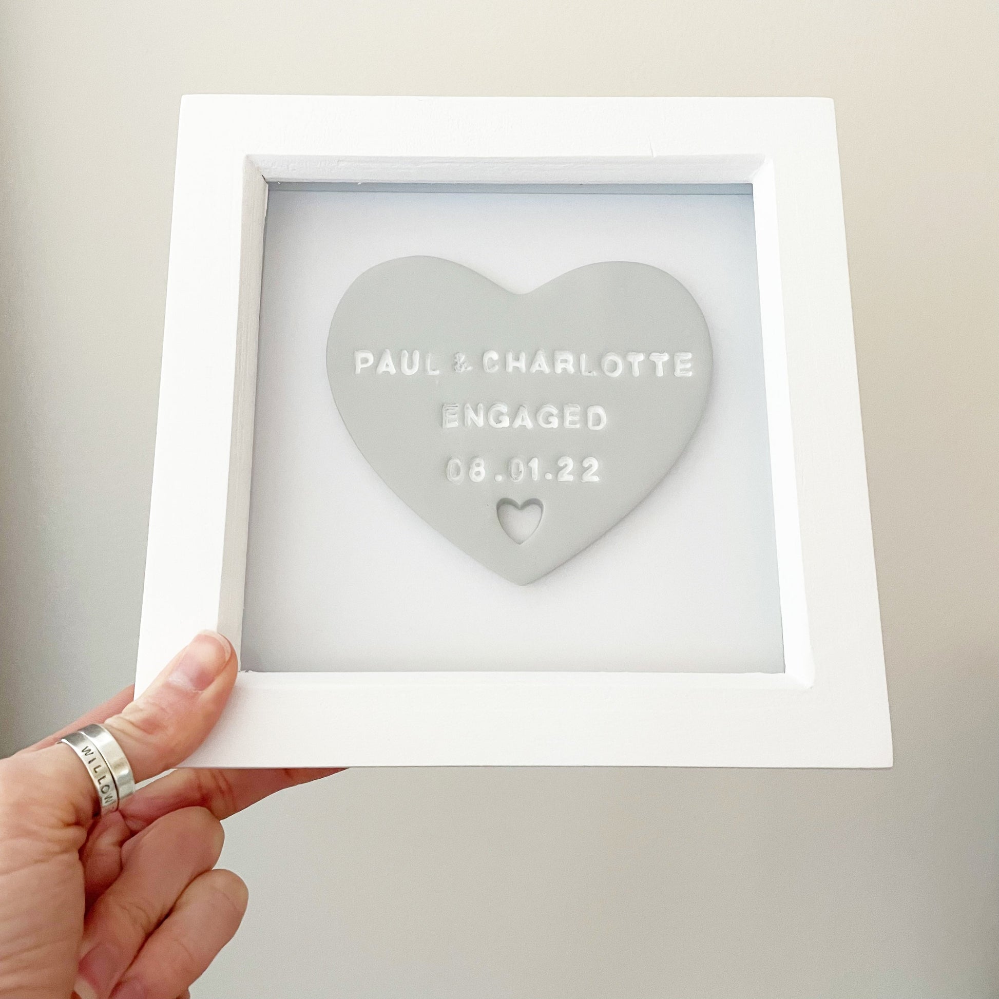 Personalised engagement gift, grey clay heart with a heart cut out at the bottom in a white box frame, the heart is personalised with PAUL & CHARLOTTE ENGAGED 08.01.22
