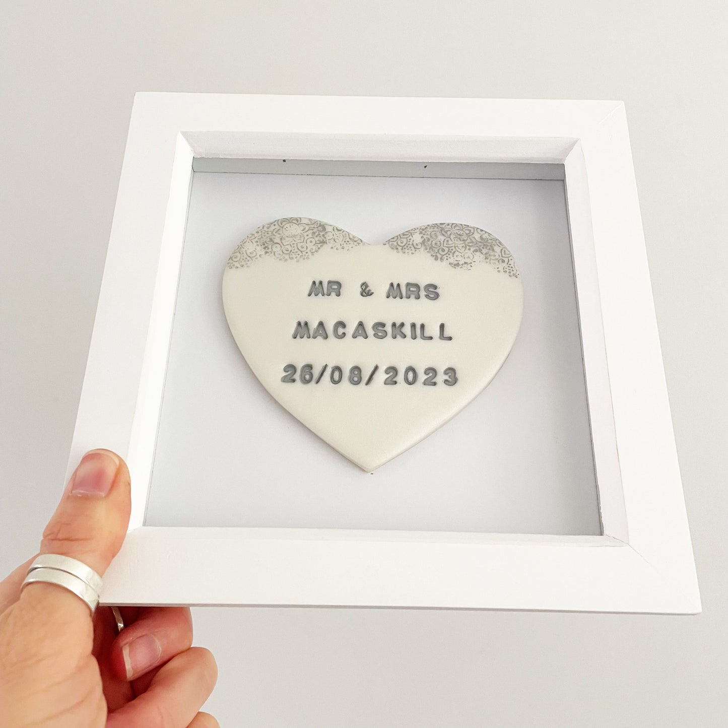 Personalised framed wedding gift, pearlised white clay heart with a grey lace edge at the top of the heart in a white box frame, the heart is personalised with MR & MRS MACASKILL 26/08/2023