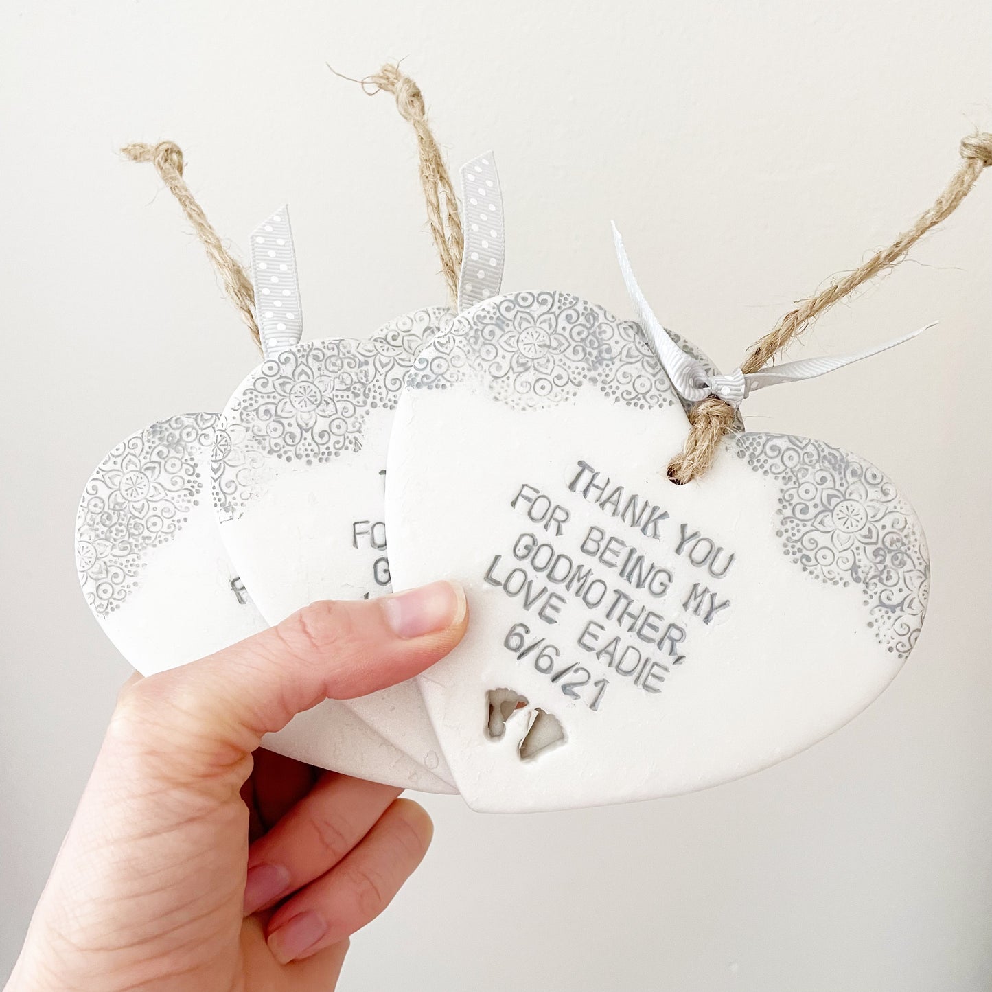 Personalised Godmother gift, 3 pearlised white clay hanging heart with baby feet cut out of the bottom and grey lace edge at the top of the heart, the heart is personalised with THANK YOU FOR BEING MY GODMOTHER LOVE EADIE 6/6/21