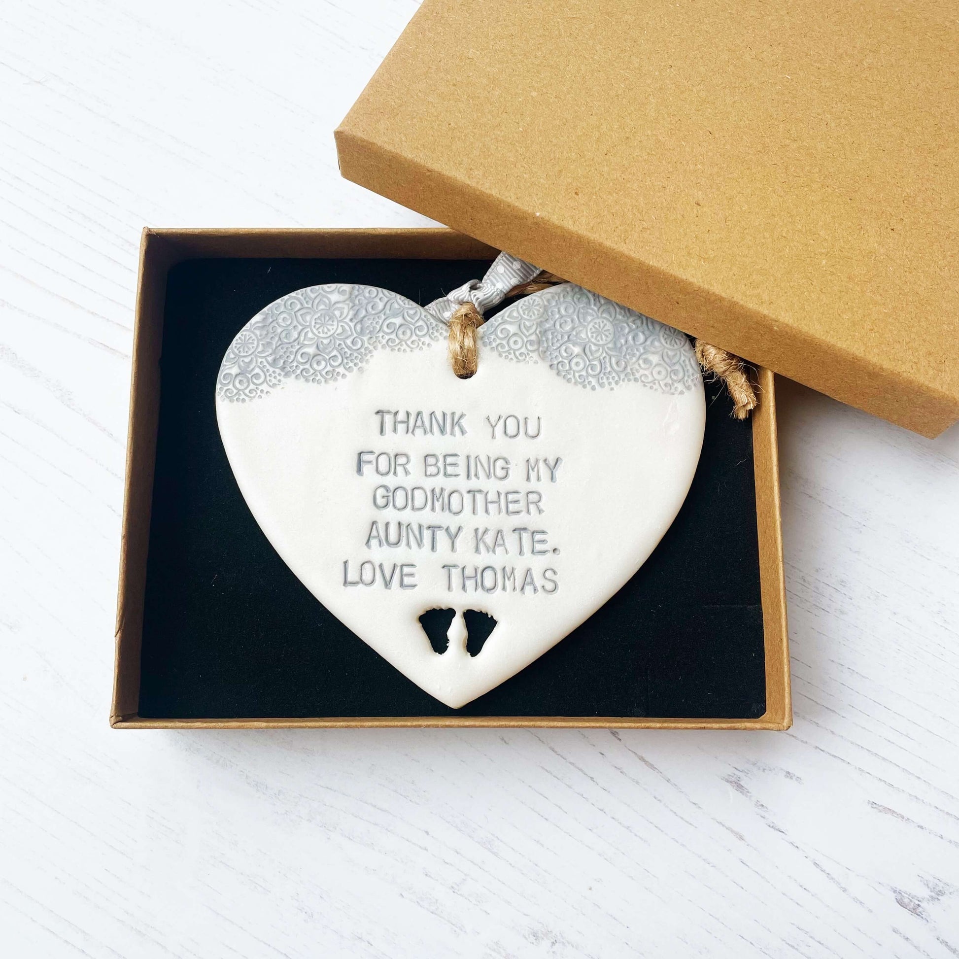 Personalised Godmother gift, pearlised white clay hanging heart with baby feet cut out of the bottom and grey lace edge at the top of the heart, the heart is personalised with THANK YOU FOR BEING MY GODMOTHER AUNTY KATE. LOVE THOMAS