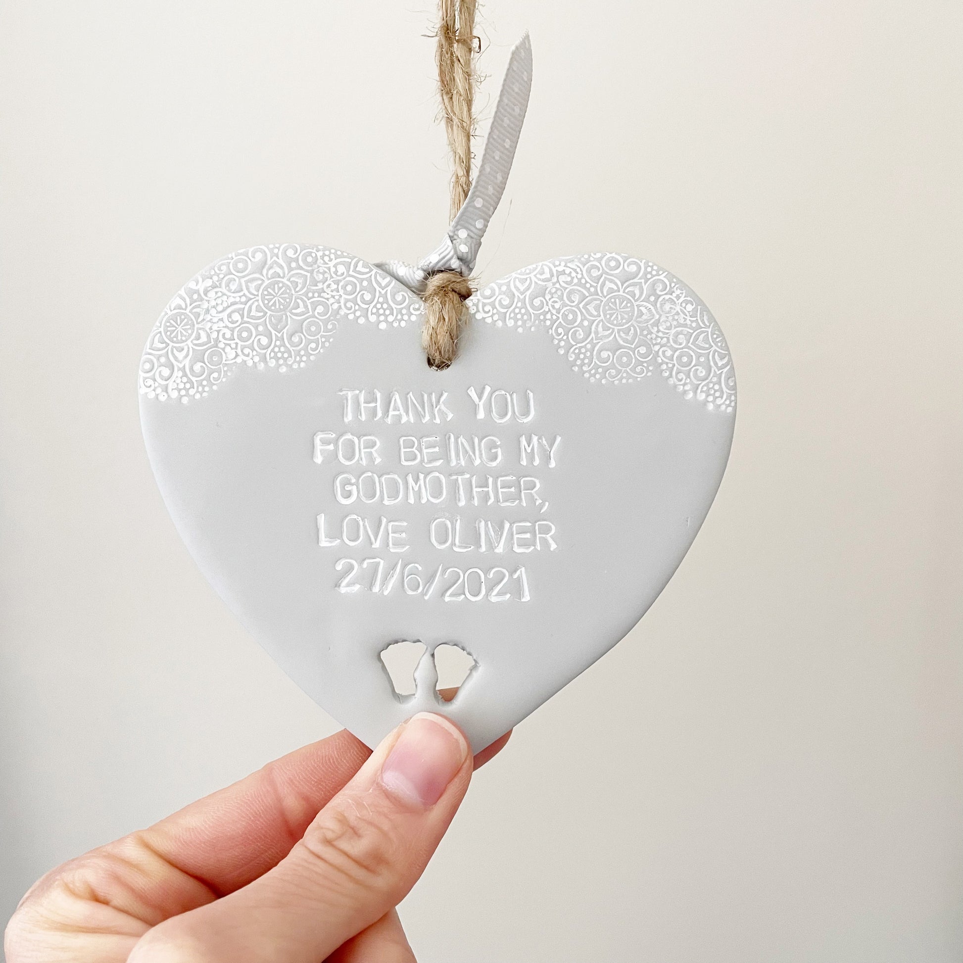 Personalised Godmother gift, grey clay hanging heart with baby feet cut out of the bottom and white lace edge at the top of the heart, the heart is personalised with THANK YOU FOR BEING MY GODMOTHER LOVE OLIVER 27/6/2021