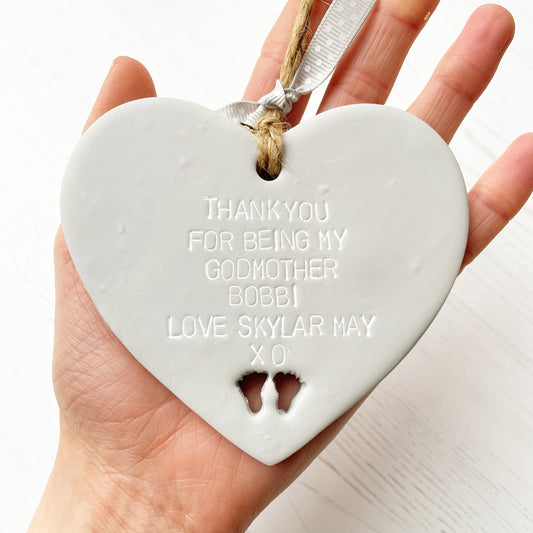 Personalised Godmother gift, grey clay hanging heart with baby feet cut out of the bottom, the heart is personalised with THANK YOU FOR BEING MY GODMOTHER BOBBI LOVE SKYLAR MAY XO