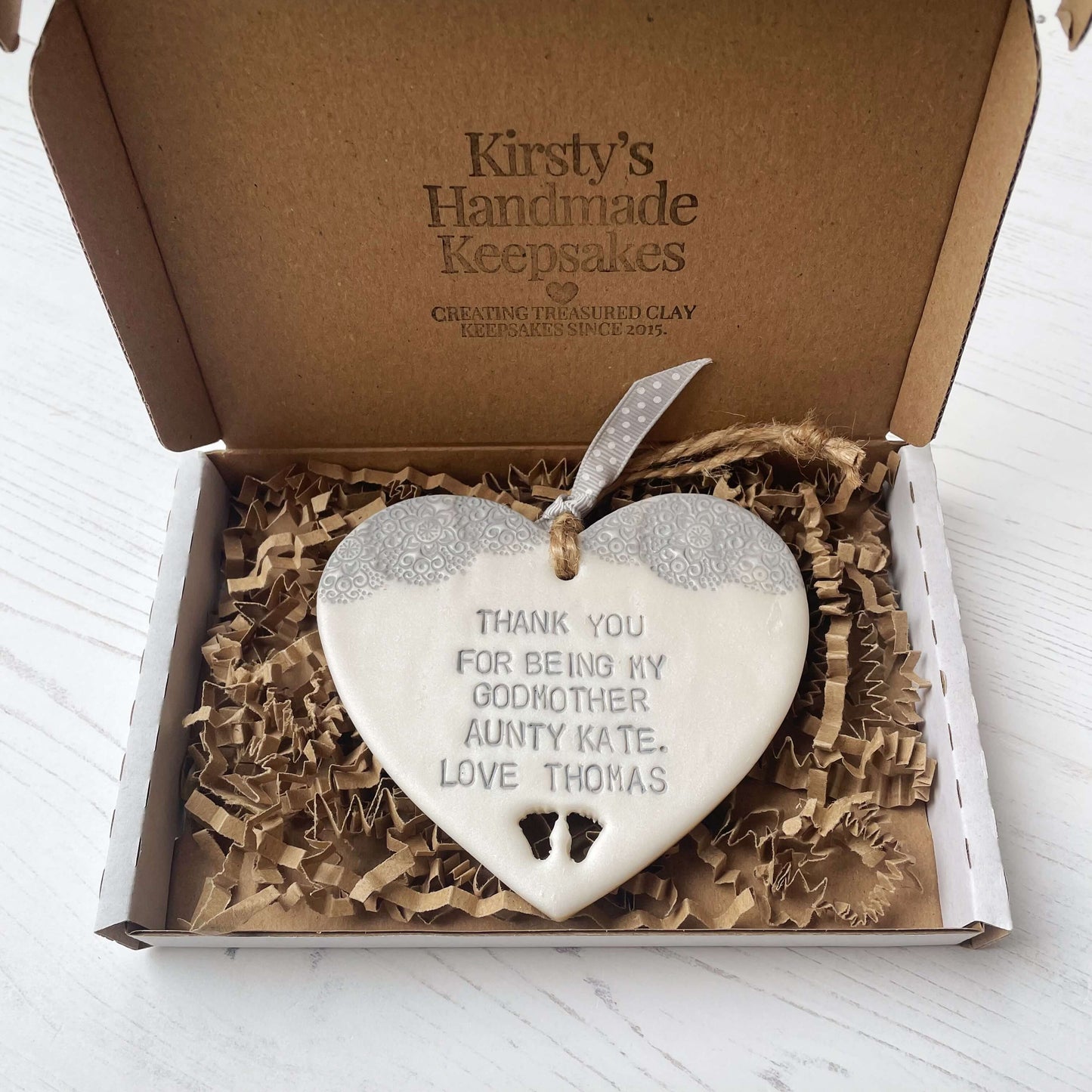 Personalised Godmother gift, pearlised white clay hanging heart with baby feet cut out of the bottom and grey lace edge at the top of the heart, the heart is personalised with THANK YOU FOR BEING MY GODMOTHER AUNTY KATE. LOVE THOMAS