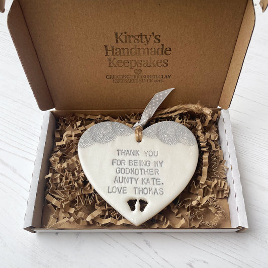 Personalised Godmother gift, pearlised white clay hanging heart with baby feet cut out of the bottom and grey lace edge at the top of the heart, the heart is personalised with THANK YOU FOR BEING MY GODMOTHER AUNTY KATE. LOVE THOMAS In a postal box with brown Kraft shredded zigzag paper