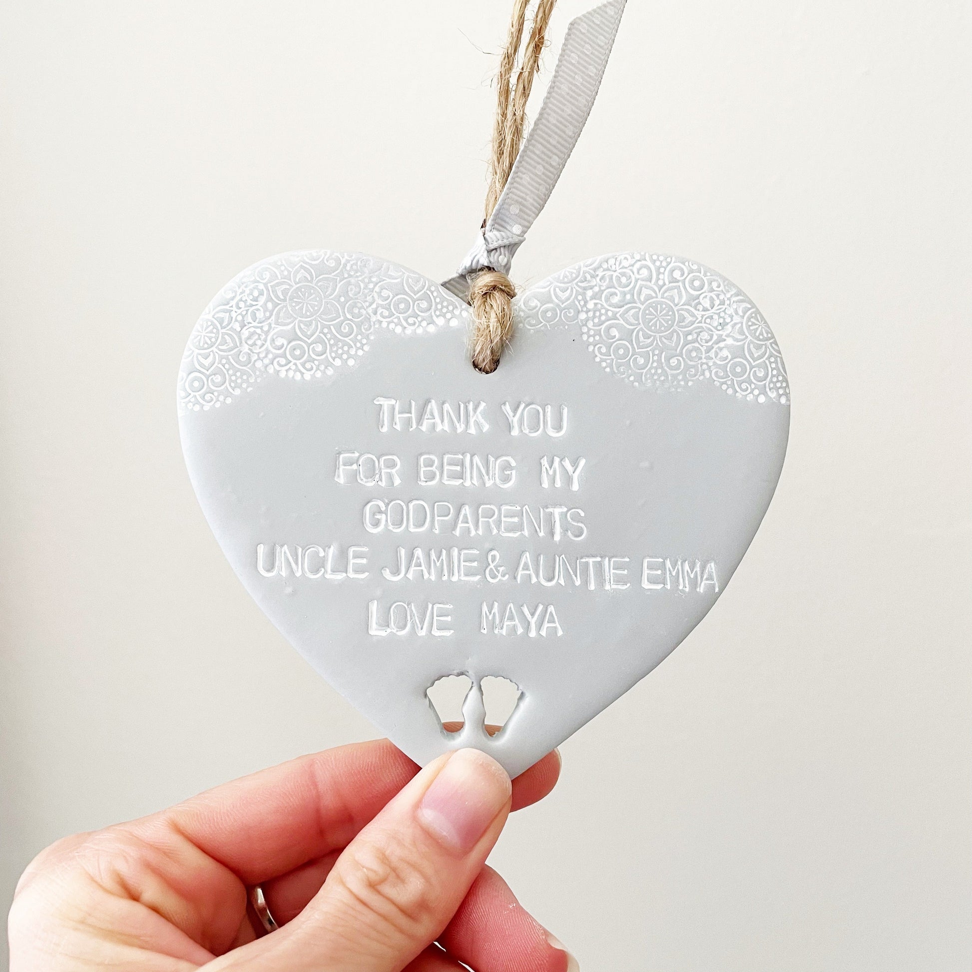 Personalised Godparent gift, grey clay hanging heart with baby feet cut out of the bottom and white lace edge at the top of the heart, the heart is personalised with THANK YOU FOR BEING MY GODPARENTS UNCLE JAMIE & AUNTIE EMMA LOVE MAYA